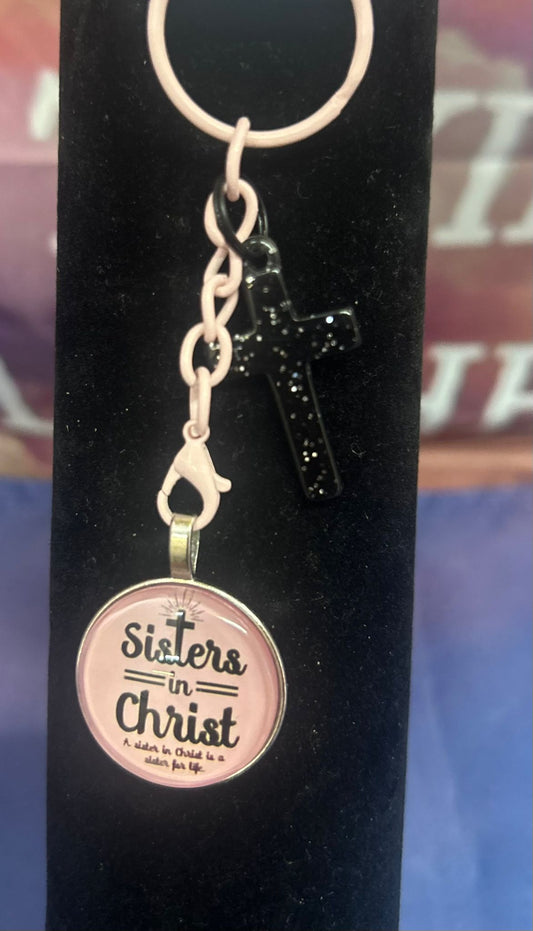 Elegante sleutelhanger van Onlyheknows2025, genaamd 'Stijlvolle Sleutelhanger Sisters In Christ', met een glanzend zwart kruisje en een ronde hanger met de inscriptie 'Sisters in Christ are sisters for life', perfect voor een waardevol geschenk.